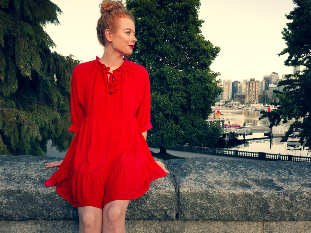 Paula sitting on a stone wall in a bright red short wilder gown with Vancouver's Coal Harbor in the background