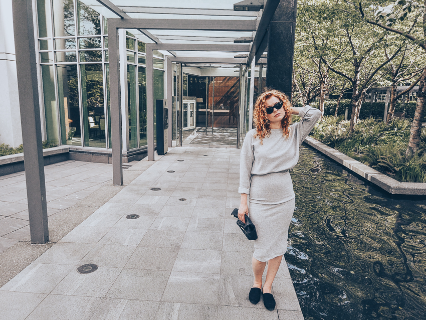 Paula posing outside a windowed apartment with her hand up to her hair in grey matching separates made with the LB Pullover and a knit jersey skirt. 